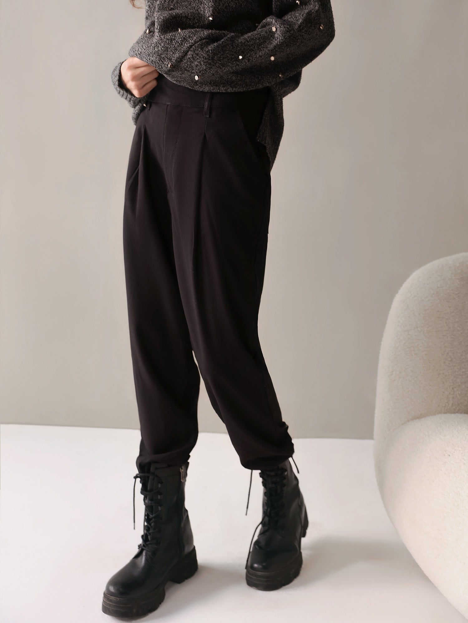 A person standing wearing black high-waisted, baggy fit pants with pleats on the front, cross pockets on the sides, and an extended waist belt. They are also wearing black boots and a sweater.