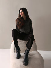 A person standing wearing black high-waisted, baggy fit pants with pleats on the front, cross pockets on the sides, and an extended waist belt. They are also wearing black boots and a sweater.