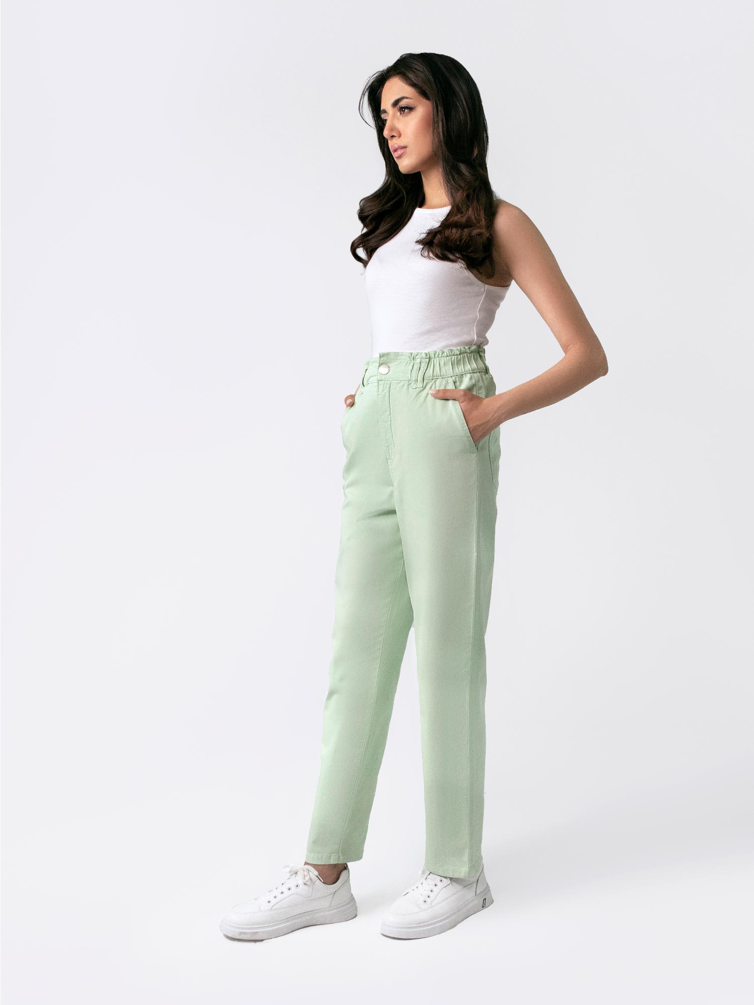 A female model wearing casual wear sage green paper bag pants with a loose fit and elasticated waist, featuring front cross pockets. She is also wearing a white top with 'Saint' printed on it and white sneakers.