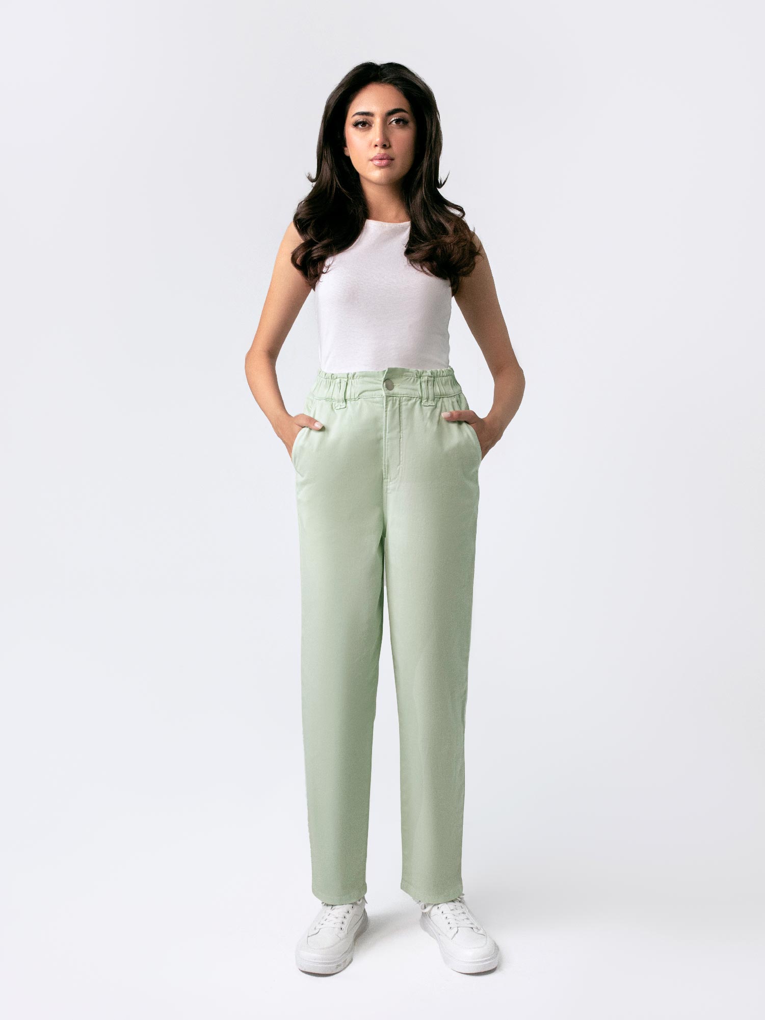 A female model wearing casual wear sage green paper bag pants with a loose fit and elasticated waist, featuring front cross pockets. She is also wearing a white top with 'Saint' printed on it and white sneakers.