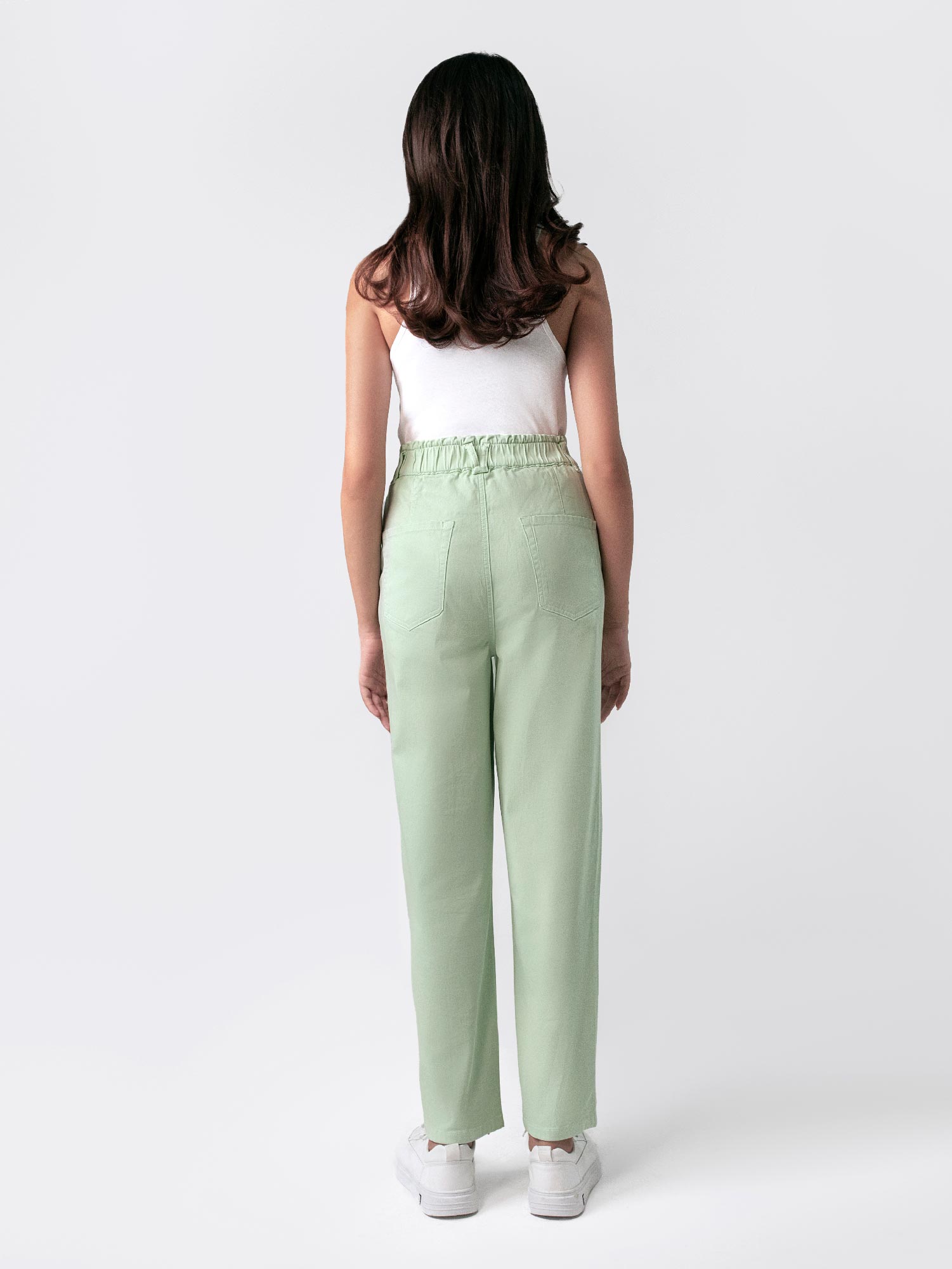 A female model wearing casual wear sage green paper bag pants with a loose fit and elasticated waist, featuring front cross pockets. She is also wearing a white top with 'Saint' printed on it and white sneakers.