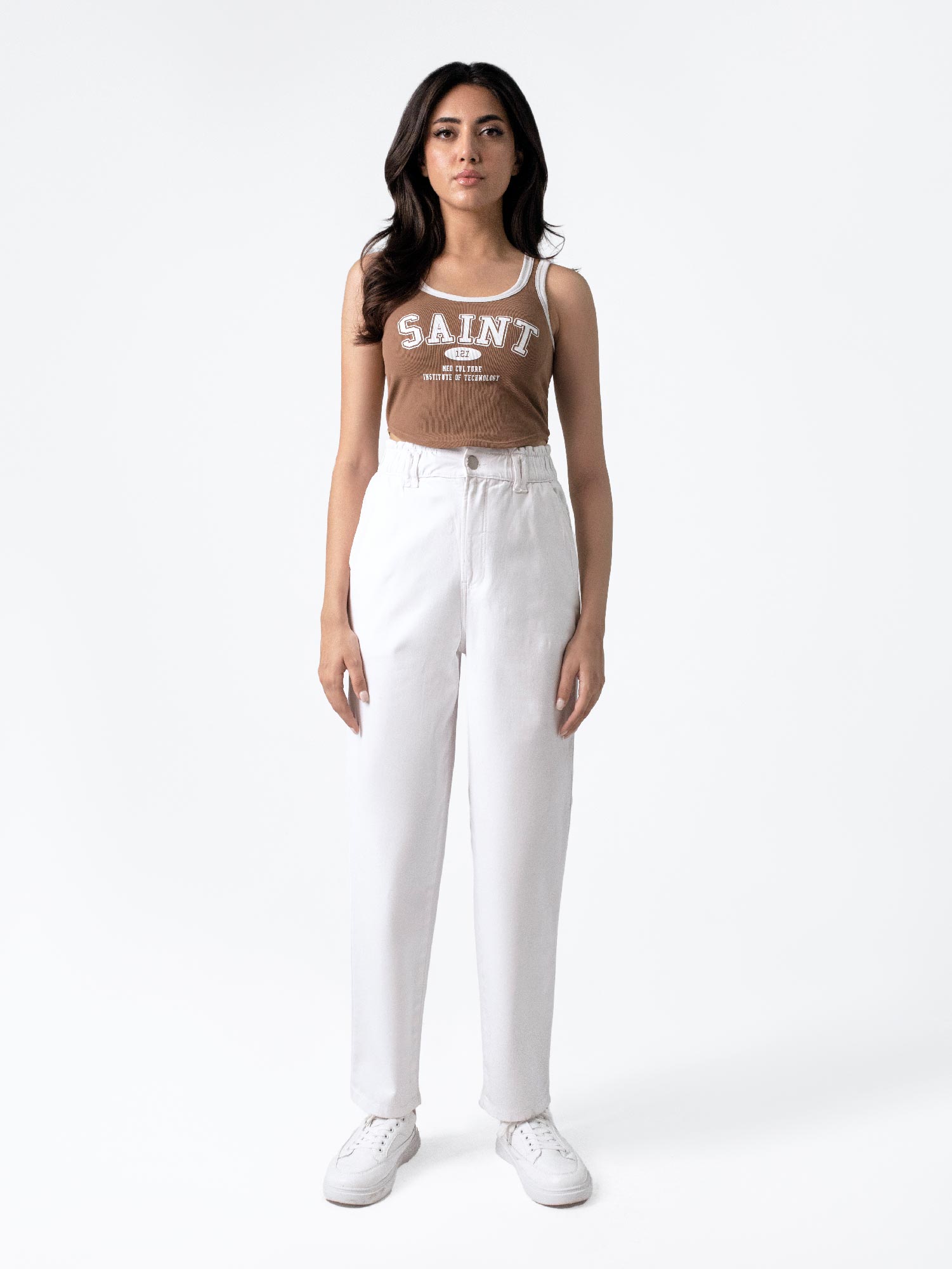 A female model wearing casual wear white paper bag pants with a loose fit and elasticated waist, featuring front cross pockets and back patch pockets. She is also wearing a brown top with 'Saint' printed on it and white sneakers.
