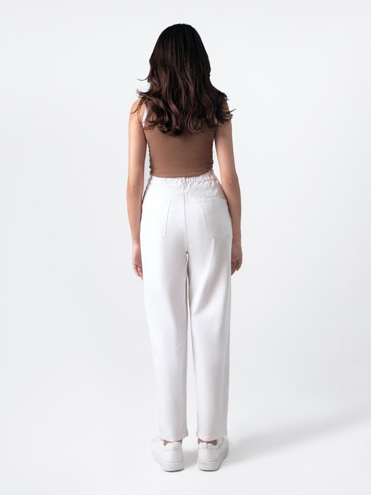 A female model wearing casual wear white paper bag pants with a loose fit and elasticated waist, featuring front cross pockets and back patch pockets. She is also wearing a brown top with 'Saint' printed on it and white sneakers.